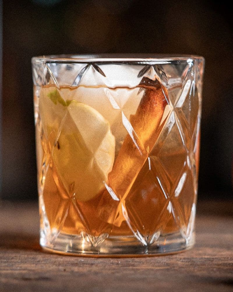 Maple-finished bourbon and spiced rum being poured for an Apple Crisp build, warm tungsten light catching the glass.