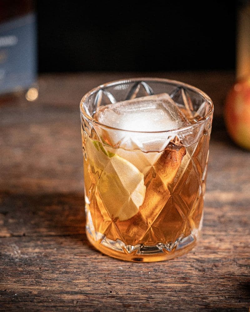 Apple Crisp cocktail set on a candlelit bar with apple slices and cinnamon sticks at hand.