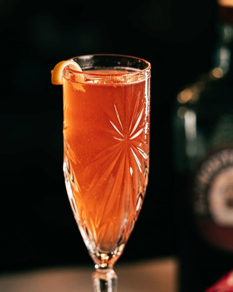 Two flutes of Jewels of Winter on a candlelit table with lemon peels and a bowl of fresh fruit.